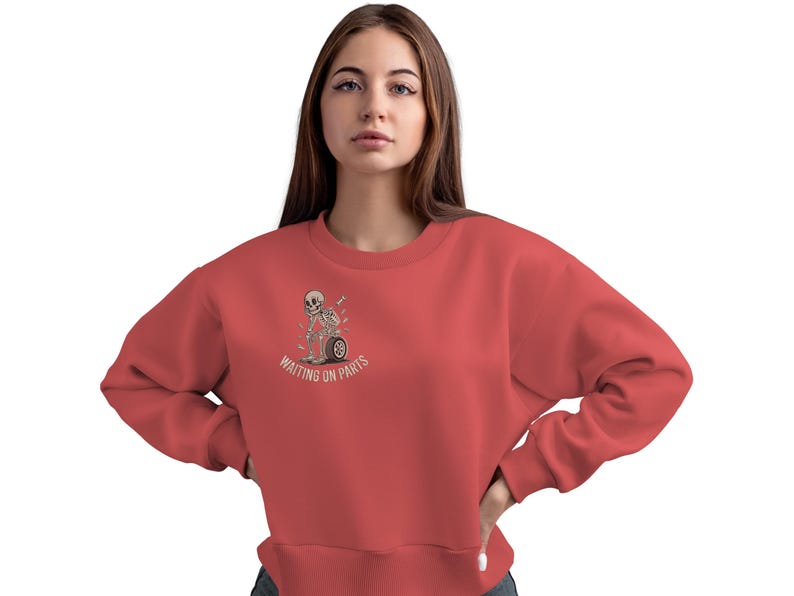 May include: Coral-colored sweatshirt featuring a graphic of a skeleton sitting on a motorcycle wheel, with the text "WAITING ON PARTS." The crew neck sweatshirt is a casual style.