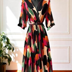 Könnte beinhalten: Ein farbenfrohes, gemustertes Kleid mit V-Ausschnitt und plissiertem Rock. Das Kleid hat einen schwarzen Gürtel und kurze Ärmel. Das Design umfasst orange, rosa, grüne und schwarze abstrakte Formen. Das Kleid wird auf einer schwarzen Schaufensterpuppe präsentiert.