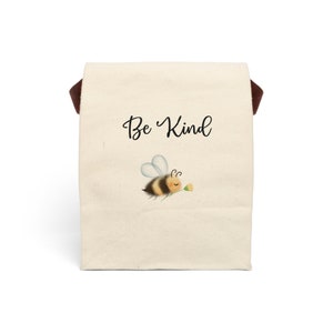 May include: A beige tote bag with a brown handle. The bag features the words "Be Kind" in a script font, along with an illustration of a cartoon bee carrying a flower. The bee has yellow and black stripes.