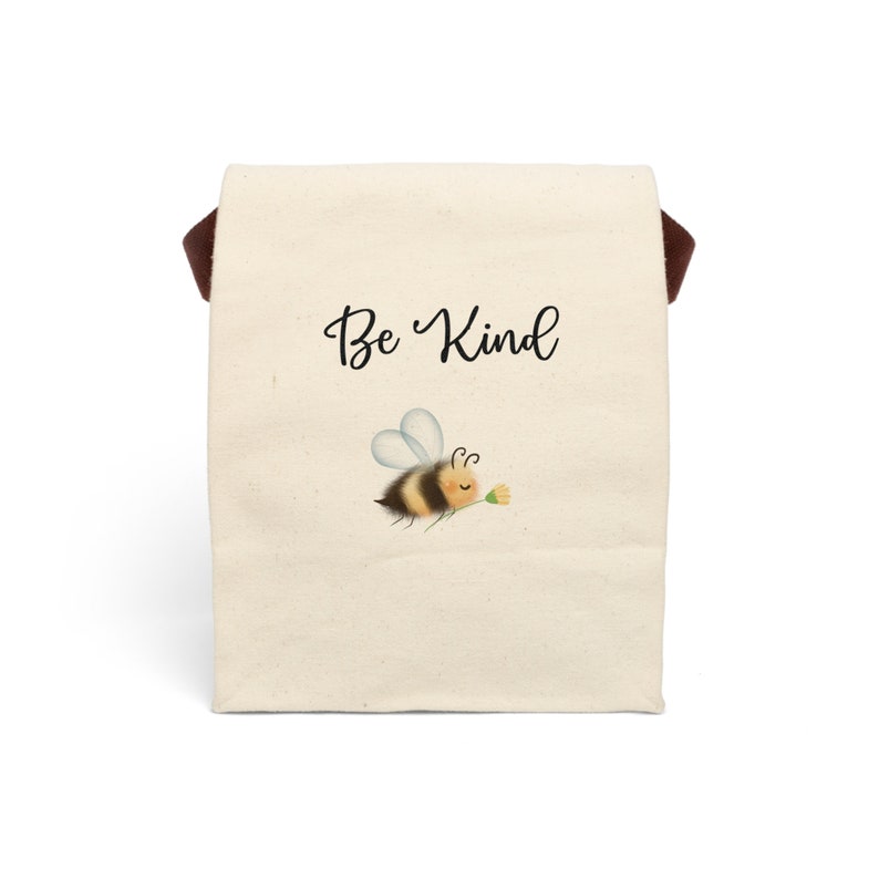 May include: A beige tote bag with a brown handle. The bag features the words "Be Kind" in a script font, along with an illustration of a cartoon bee carrying a flower. The bee has yellow and black stripes.