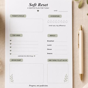 Może przedstawiać: Biały planer z napisem "Soft Reset" i "A Gentle Plan for Today". Zawiera sekcje dotyczące skupienia, sukcesów, posiłków i nawodnienia. Z prawej strony planera leży złoty i biały długopis.