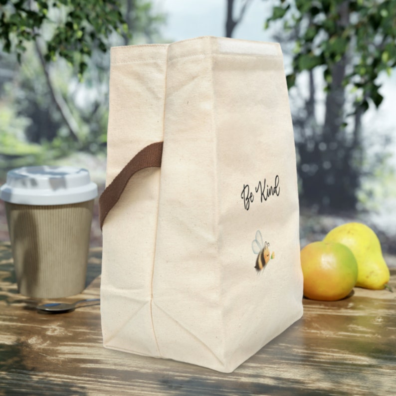 May include: A beige canvas lunch bag with a brown handle and the words "Be Kind" printed on the front, along with a cartoon bee. A disposable coffee cup, an apple, and a pear are on a wooden surface.