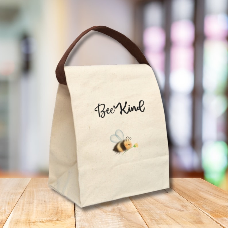 May include: A beige canvas lunch bag with a brown handle. The bag features the words "Bee Kind" in black script, along with an illustration of a cartoon bee holding a flower. The bag is designed to carry food and drinks.