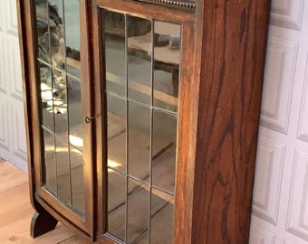 May include: A dark brown wooden wall-mounted cabinet featuring two glass doors with a grid pattern of leaded cames. The cabinet has a curved top crest with a central circular detail and a small shelf on top, showcasing a visible wood grain.