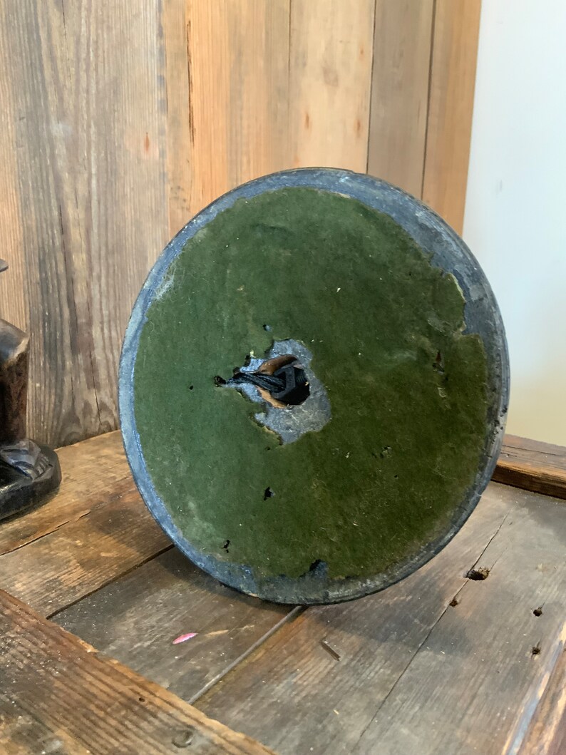 May include: The bottom of a round, dark gray lamp base with a green felt covering. The felt is worn and has a hole in the center.