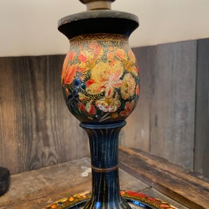 May include: A black wooden lamp base with a floral design painted in shades of orange, yellow, and green. The base has a round, decorative tray at the bottom.