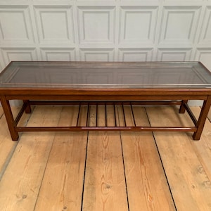 May include: A rectangular coffee table constructed from medium-toned wood featuring a clear glass top insert. The table has four straight legs and a lower slatted shelf for storage, providing a flat surface area for placement of various household items.