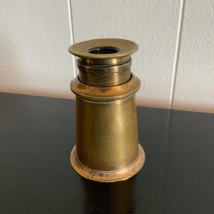 Puede incluir: Un telescopio antiguo de latón. Tiene una base acampanada y un cuerpo cilíndrico con anillos roscados y un ocular circular. El latón tiene una pátina cálida y envejecida. El telescopio está diseñado para ver objetos distantes. Altura aproximada 10 cm.