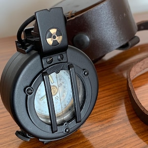 May include: A black compass with a gold and black symbol on top, a light blue and white compass dial, and a brown leather case. The compass has a leather strap and is set on a wooden surface.