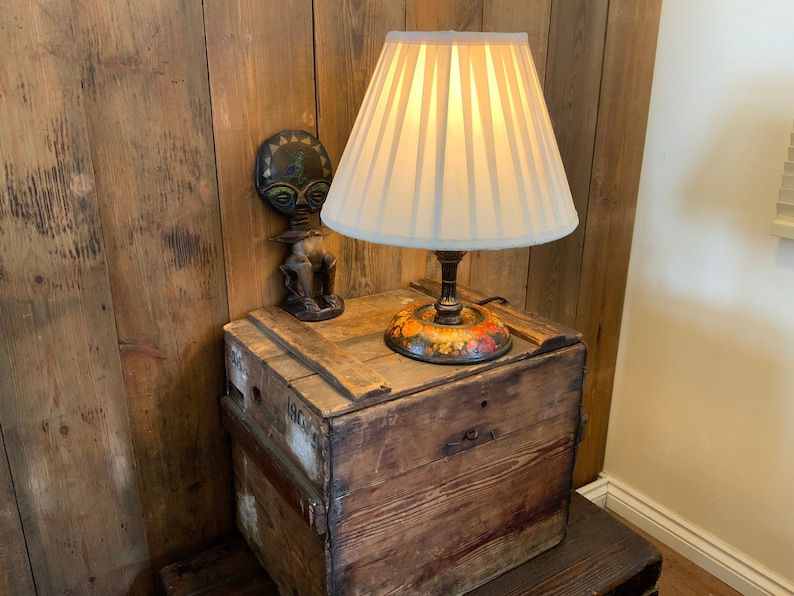 May include: A white lampshade with pleated fabric sits atop a wooden base with a colorful floral design. The lamp is on a wooden crate with a small wooden figure with a round head and a black and green design.