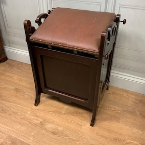 Könnte beinhalten: Ein dunkelbrauner Holzhocker mit einem braunen Ledersitz. Der Hocker hat ein Staufach mit einer Tür und dekorativen Schnitzereien. Der Hocker steht auf einem Holzboden.