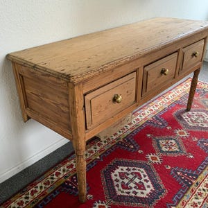 May include: A light brown wooden console table with three drawers and brass knobs. The table has a distressed finish and is supported by four slender legs. The table is placed on a red patterned rug.