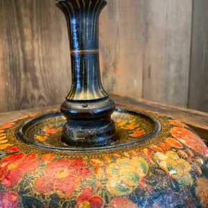 May include: A black lamp base with a floral design. The base is made of wood and has a round, flat top. The floral design is painted in shades of red, yellow, and green.