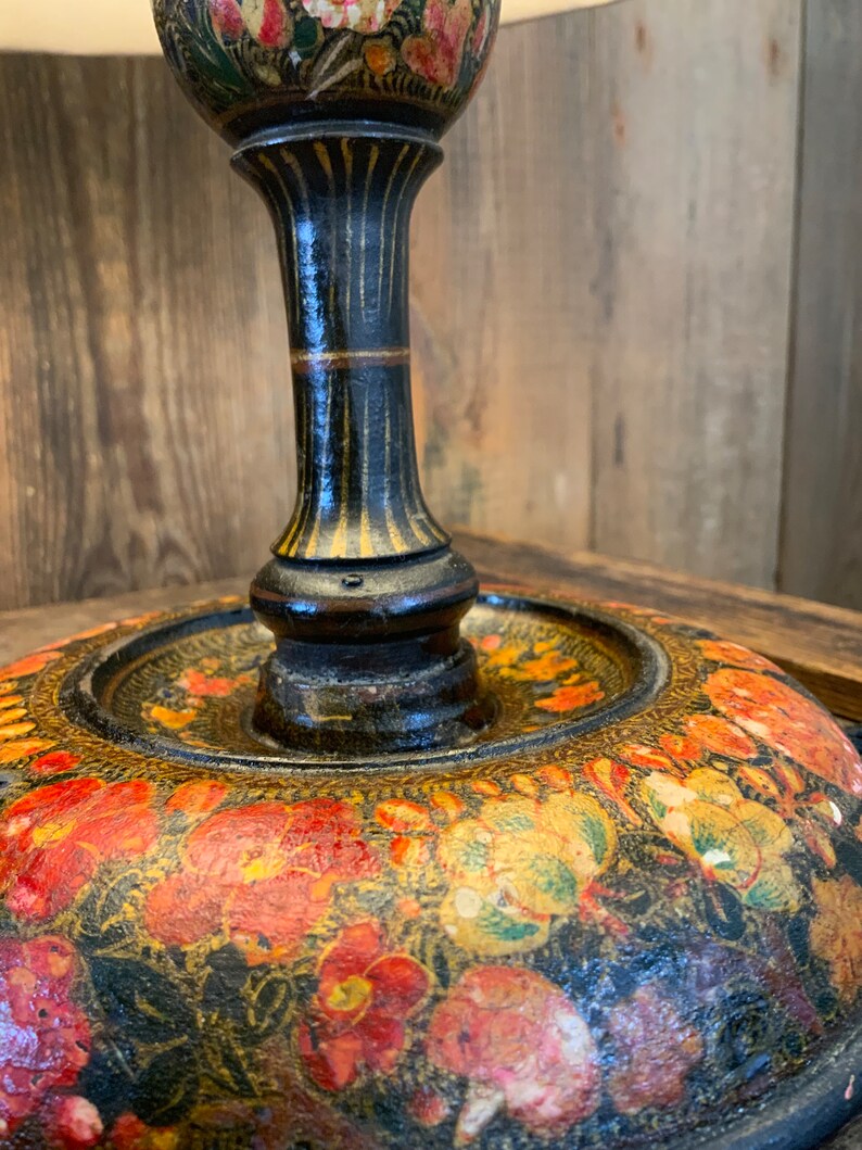 May include: A black lamp base with a floral design. The base is made of wood and has a round, flat top. The floral design is painted in shades of red, yellow, and green.