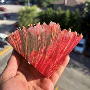 May include: A triangular piece of stabilized wood featuring a vibrant red and natural wood grain pattern. The top edge has a jagged, raw, and uneven texture, while the flat surfaces display a polished finish with visible wood fibers and streaks.