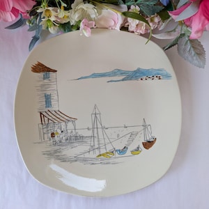 May include: A square off-white ceramic plate featuring a line-art illustration of a coastal scene. The design includes a multi-storey building, small boats in blue, yellow, and brown, and distant mountains rendered in blue ink on the surface.