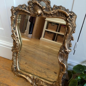May include: A decorative, rectangular mirror with an elaborate gold-coloured frame. The mirror reflects a room with wooden flooring and a white cabinet. The frame has a shell-like motif at the top.