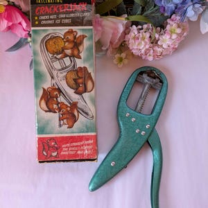 May include: Vintage Crackerjack nutcracker with original box. The box features illustrations of squirrels cracking nuts. The nutcracker is teal-coloured and has a lever design. The box text reads "Cracks Nuts, Crab & Lobster Claws, Crushes Ice Cubes."