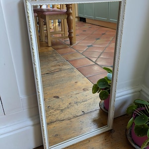 May include: A rectangular mirror with a white frame and gold trim. The mirror is leaning against a wall on a wooden floor. The reflection shows a room with a table and plants.