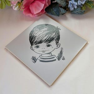 May include: A square, pale grey tile with a black and white illustration of a young boy with dark hair and a striped top. The boy's face is in the centre of the tile, with a small smile. The tile is likely ceramic or a similar material.