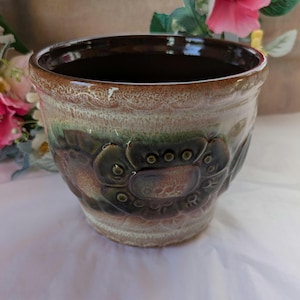 May include: A ceramic plant pot with a dark brown interior and a textured exterior. The pot features a floral design in shades of green and brown. The rim is a lighter brown colour.