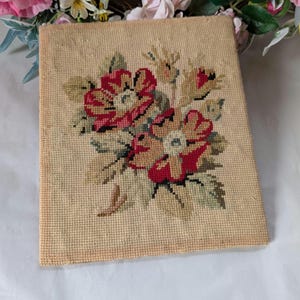 May include: A vintage needlepoint tapestry with a floral design. The artwork features a bouquet of red and peach flowers with green leaves on a beige background. The rectangular piece appears to be hand-stitched, showcasing intricate detail.