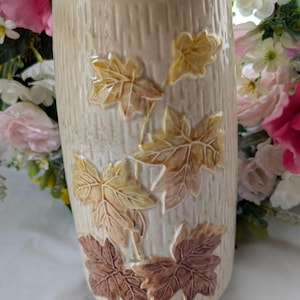 May include: A beige ceramic vase with a textured, bark-like surface. The vase is adorned with raised, three-dimensional maple leaves in shades of yellow, brown, and tan. The vase is approximately 20 cm tall.