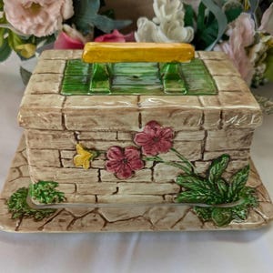May include: A ceramic butter dish with a brick design and a green and yellow bench on top. The dish is decorated with pink and yellow flowers and green leaves.