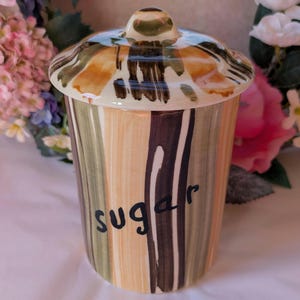 May include: A ceramic sugar jar with a lid. The jar is decorated with brown, green and yellow stripes. The word "sugar" is written in black on the front of the jar.
