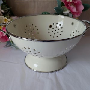 May include: A cream-coloured enamel colander featuring a wide bowl with numerous drainage holes, a pedestal base, and two silver-toned metal handles on opposite sides. The rim and handles are finished with a polished silver-toned metal trim.