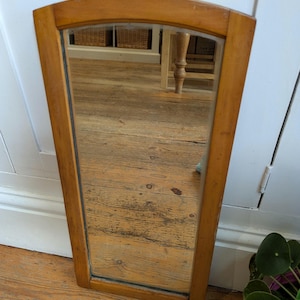 May include: A long, rectangular mirror with a light brown wooden frame and a curved top. The mirror is positioned against a white wall and a wooden floor. The reflection shows a room with baskets and a table. The mirror's height is approximately 70 cm.