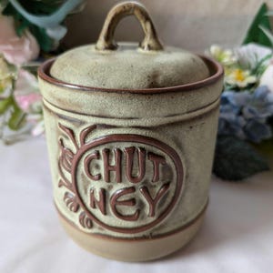 May include: A green ceramic jar with a brown lid and handle. The jar has a brown circle with the word "CHUTNEY" written in it.
