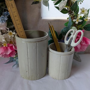 May include: Two cylindrical off-white ceramic crocks with vertical ribbed textures. A wooden ruler measuring up to 23 cm is inside the larger crock. The smaller crock holds two yellow and black pencils and a pair of white scissors.