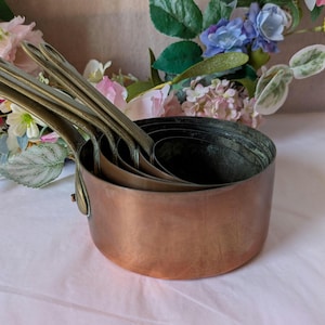 May include: A set of nested copper measuring cups with long, curved brass handles. The cups have a warm, metallic finish and are stacked inside one another, showing a graduated size from the largest outer cup to the smallest inner cup.