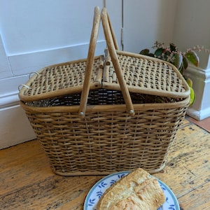 May include: A light brown wicker picnic basket with a hinged lid and wooden handle. A plate with a loaf of bread sits in front of the basket. The basket appears to be vintage.