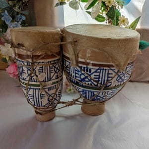 May include: A pair of connected bongo drums featuring a hand-painted white ceramic body with blue and green geometric patterns. The drum heads are made of natural tan animal skin, secured with a network of thin, light brown twine rope.