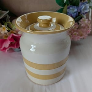 May include: A ceramic jar with a lid, featuring a white and ochre striped design. The jar has a rounded shape with a slightly flared rim and a small knob on the lid. The colour scheme is neutral, with contrasting stripes.
