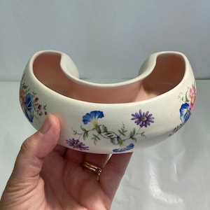 May include: A white ceramic planter shaped like a horseshoe with a pink interior. The planter is decorated with a floral pattern in shades of blue, pink, purple, and green.
