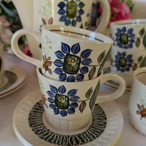 May include: A vintage cream-coloured tea set with a floral design. The set includes a teapot, cups, saucers, and a creamer. The floral pattern features blue, green, and brown flowers. The cups are stacked on a saucer with a dark green and white pattern.