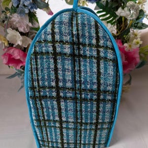 May include: A tea cosy in a blue and teal plaid pattern, edged with a light blue trim and a small loop. The fabric has a textured appearance. The background shows a floral arrangement.
