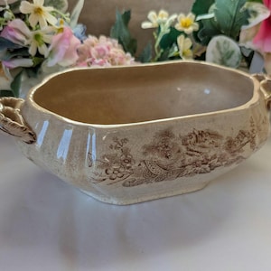 May include: A vintage, cream-coloured ceramic serving bowl with a brown landscape pattern. The bowl has a rectangular shape with rounded corners and ornate handles on each side. The interior is a light brown colour.