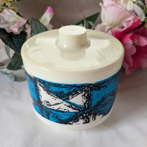 May include: A vintage ceramic container in white and blue, with a lid and a small handle. The container is decorated with a geometric pattern in blue and black. Ideal for kitchen storage or home decor.