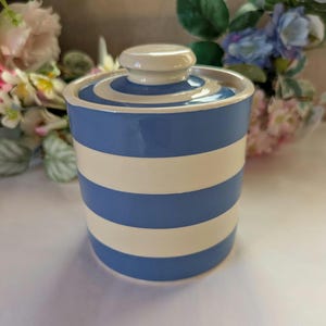 May include: A cylindrical ceramic canister featuring a repeating pattern of horizontal blue and white stripes. The container includes a matching lid with a central knob handle, both decorated with the same blue and white striped design.