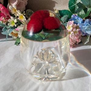 May include: A clear glass paperweight with a detailed, flower-like design at the base. The top features three realistic red raspberries with green leaves, encased in clear resin. Decorative item.