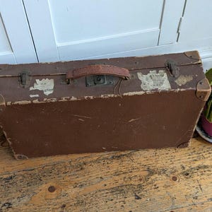 May include: A rectangular brown vintage suitcase featuring a worn leather handle, metal corner reinforcements, and two metal latches. The exterior surface shows signs of age with faded paper labels and scuff marks across the textured material.