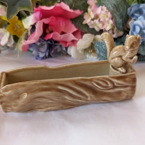 May include: A ceramic planter shaped like a tree trunk with a squirrel figurine on top. The planter is a light brown colour and has a textured surface.