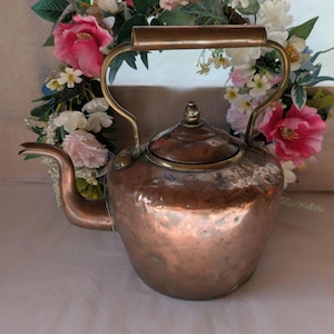 May include: A copper kettle with a brass handle and spout. The kettle has a rounded body and a lid with a small knob. The handle is made of wood. The kettle is likely vintage or antique.