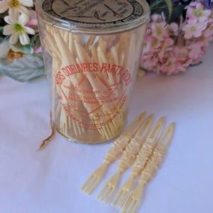 May include: A clear plastic container filled with off-white plastic hors d'oeuvres party picks. The container has a red and black label. Four picks are outside the container, showing a decorative design.