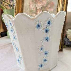 May include: White ceramic planter with a scalloped rim and blue floral design. The planter has a hexagonal shape and is decorated with delicate blue flowers.