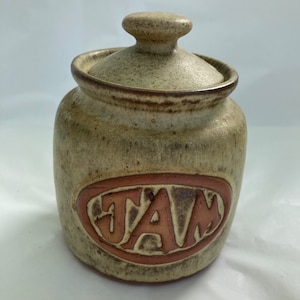 May include: A ceramic jar with a lid, decorated with a brown glaze and the word "JAM" in a red oval.
